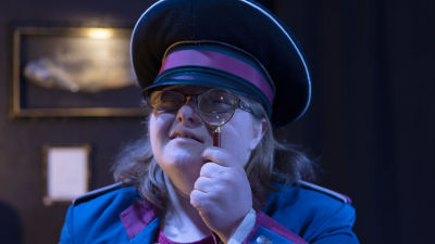 En person klädd som en museivakt kikar mot kameran genom ett förstoringsglas.