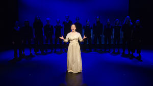 De historiska scenerna varvas med stämningsfulla livemusiknummer. En kvinna i vit klänning sjunger på en scen. Bakom henne står en grupp svartklädda gestalter i en halvcirkel.
