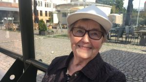 Att man kan åka två timmar på en biljett, det tycker pensionären Hilkka Halonen är underbart. Då hinner man med mycket, säger hon. äldre kvinna med vit keps sitter i busskuren och väntar på en buss och ler mot kameran