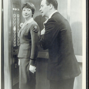 Hississä hymyilevä nainen katsoo olan yli miestä korjaamassa solmiotaan ja puhumassa hänen takanaan.