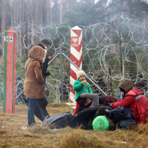 En grupp människor står och ligger på marken vid ett taggtrådsstängsel. Vid stängslet står Belarus röd-gröna och Polens röd-vita gränspålar mittemot varandra.