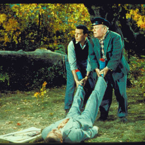 Kaksi miestä, toinen iäkäs ja toinen nuori, pitävät kiinni maassa tiedottomana makaavan miehen jaloista ja katsovat jonnekin sivullepäin. Kuva elokuvasta Mutta kuka murhasi Harryn?