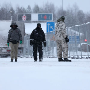 Toppavaatteisiin pukeutunut tummaihoinen ihminen kävelee poliisin saattamana metallisen aidan reunusta lumisateessa, aidan vieressä maastopukuisia varusmiehiä.