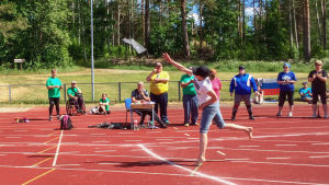 Urheilukentällä nainen heittää saapasta, ympärillä ihmisiä katsomassa, taustalla puita