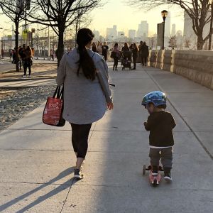 Mycket hänger på kvinnorna i USAs barnfamiljer. 3-4-årig pojke på sparkbräda på trottoar i solnedgång, Manhattan i bakgrunden