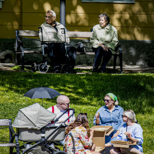 En äldre kvinna och en äldre man sitter på en parkbänk, en familj sitter i gräset och äter pizza en bit bort.