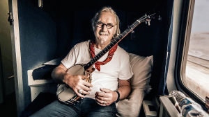 Kiehtova maailma: Billy Connollyn Amerikan seikkailu. Mies istuu junassa kitara kädessä ja katsoo kameraan.