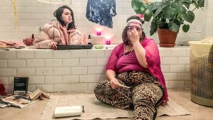 Kaksi naista kylpyhuoneessa. Toinen istuu kylpyammeessa ja toinen kylpyammeen edessä. Molemmilla on vaatteet päällä, toisella tupakka suussa.