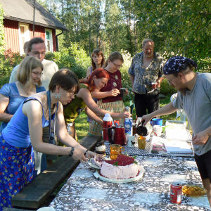 Kesäisessä puutarhassa pitkän pöydän äärellä joukko ihmisiä jonottaa vadelmakermakakkua.