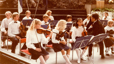 En barnorkester som spelar stråkinstrument sitter på stolar med noter framför sig. En dirigent i mörk kostym lutar sig över en flicka.