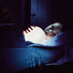En liten flicka ligger på en säng i ett mörkt rum med en lampa i famnen. 
