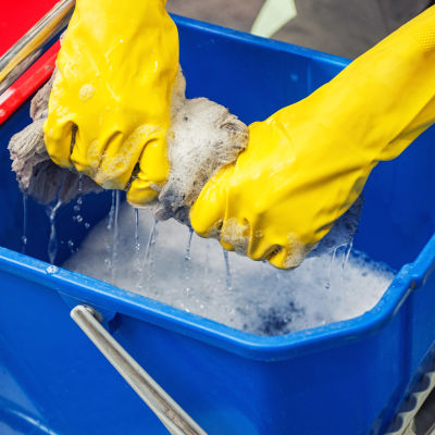 Två händer i gula gummihandskar vrider vatten ur en luddig golvmopp ner i en blå hink.
