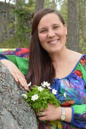 Laura Leppänen, en dam med färggrann klänning och en bukett vitsippor i handen, lutar sig mot en sten.