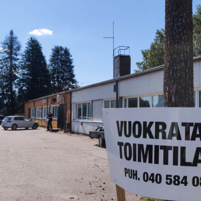 I högra hörnet på bilden syns en skylt där det står på finska att affärsutrymmen uthyres. Bkom skylten synns ett lågt långt hus.
