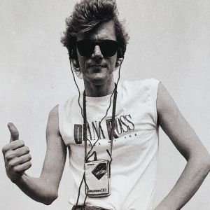 Mustavalkoisessa kuvassa 1980-luvulta mies aurinkolaseissa ja korvalappustereot korvilla näyttää peukaloa.