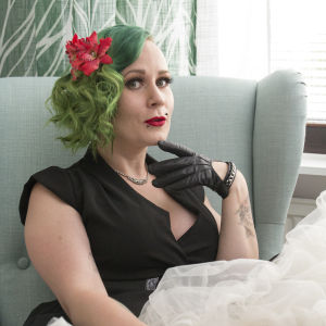 Isa älskar att dansa, och burlesk är en dansform. En kvinna med grönt hår och rött läppstift, iklädd en svart klänning, sitter på en ljusgrön fåtölj.