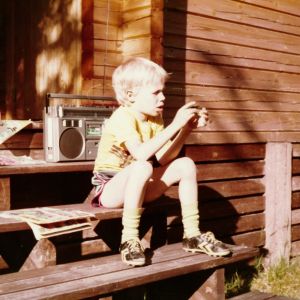 Jouni Järvelä istuu alle kymmenvuotiaana mökin portailla sarjakuvalehden ja kasettinauhurin kanssa.