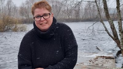 Kvinna i mörk tröja sitter på en brygga vid ett vattendrag.