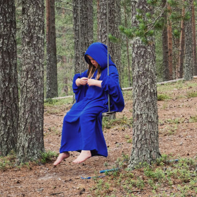 Siniseen kaapuun pukeutunut tyttö istuu keinussa metsässä.