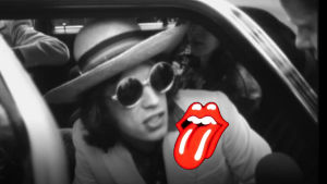 Mick Jagger sitter i bil och intervjuas 1970 i Finland. Svartvit bild med röd Rolling Stones logo på Micks vita kavaj.