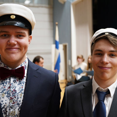 Två unga män med studentmössor. I bakgrunden skymtar Finlands flagga. De är i en gymnastiksal.