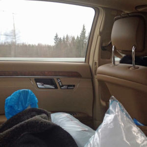 Auton takapenkillä otettu kuva niin että siniseen sairaalatossuun puettu jalka näkyy tyynyjen päällä, vasemman puoleinen sivu-ikkuna taustalla. Vanhempi nainen ajaa autoa.