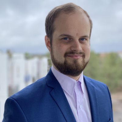 En man med brunt hår och skägg klädd i mörkblå kavaj och skjorta tittar in i kameran och ler.