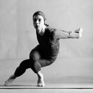 Merce Cunningham Tanssija Merce Cunningham liikkeessä, polvet ristissä, kädet selän taakse ojennettuina, yllään neulottu asu