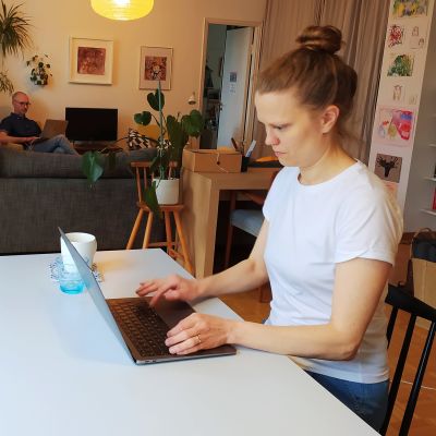 Kvinna i förgrunden skriver på bärbar dator, man i bakgrunden jobbar också på dator sittandes i en fåtölj.