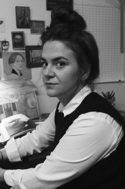 På den svartvita bilden syns illustratören Elin Sandström sitta vid ett skrivbord. Hon har håret uppsatt i en hög knut och har vit skjorta och mörk pullover på sig. På väggen bakom henne finns flera bilder bland annat syns Helene Schjerfbeck. 