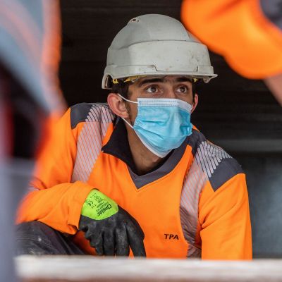 En person i hjälm och annan utrustning på ett varv jobbar. Han bär munskydd.