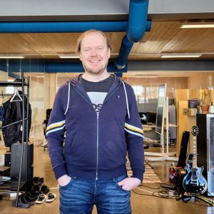 En man i blå huvtröja står med armarna i fickorna och ler mot kameran.