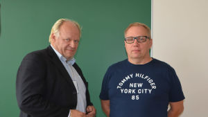 Akilles Bandys ordförande Jari Kettunen och viceordförande Kari Janhunen. Två män står framför grön vägg.