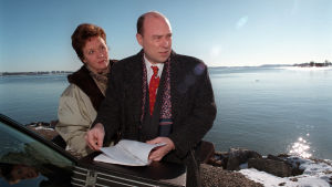 Liisamaija Laaksonen ja Pertti Sveholm näyttelivät sarjassa Presidentin mies. Mies ja nainen meren rannalla, aurinkoinen kevätsää. Mies selaa papereita, molemmat synkän oloisia.