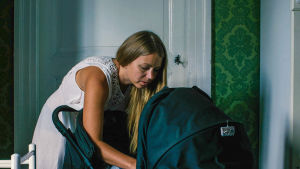 Huvudpersonens berättelse är ord för ord återgiven i dokumentären. Kvinna pysslar om barn i barnvagn inne i sitt hem.
