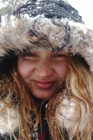 Lähikuva vaaleahiuksisesta hymyilevästä naisesta. Naisella on karvareunusteinen huppu päässä, joka on lumen kuorruttama. myös hiuksissa on lunta.