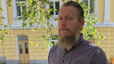 En man med grått hår och långt grått skägg står ute. I bakgrunden en stor gul byggnad, gamla stadshuset i Ekenäs. Sommar.