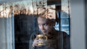 Some Deep Story. Nainen ikkunan takaa kuvattuna, kahvikuppi kädessä katsoo ulospäin.