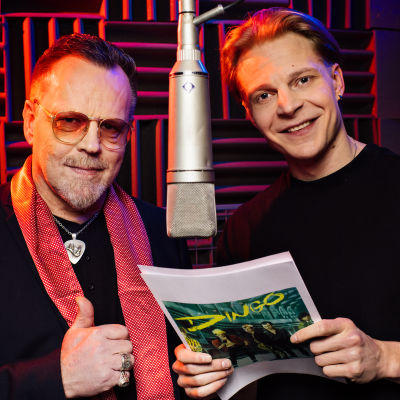 Pertti Neumann ja Olli Riipinen seisovat molemmin puolin katosta roikkuvaa mikkiä äänitysstudiossa hymyillen.