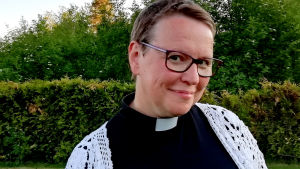 Camilla Svevar började studera till präst som 43-åring, efter ett liv som sångerska och kantor. En korthårig kvinna i närbild ler mot kameran. Kvinnan har prästskjorta på sig och en vit virkad tröja. I bakgrunden gröna träd och en häck.