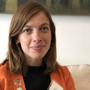 Li Andersson i New York. Porträttfoto av Li Andersson i New York, i orange kavaj med namnbricka på uppslaget.