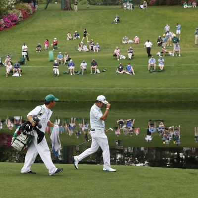 Hideki Matsuyama kävelee US Mastersissa lammen rannalla caddiensa kanssa. Lammen toisella puolella oleva yleisö heijastuu veden pinnasta.