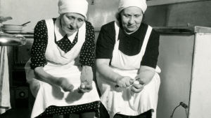 Före halvfabrikatens och frysboxarnas tid fanns det otaliga arbetsuppgifter i ett restaurangkök. De här kvinnorna arbetade i restaurangköket på Kuopio station på 1950-talet. Två kvinnor i vita förkläden och vita huvuddukar sitter och skalar potatis i restaurangköket till Kuopio station.