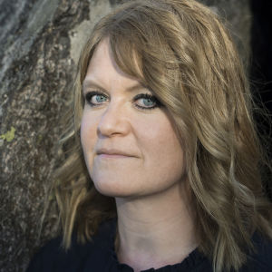 "Jaktlaget" är Ulrika Hanssons debutroman utgiven av Schildts & Söderströms. En kvinna med svart eyeliner står framför en trädstam och ser in i kameran.