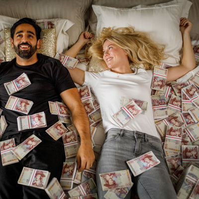 En man och en kvinna ligger på en säng med en massa sedelbuntar. 