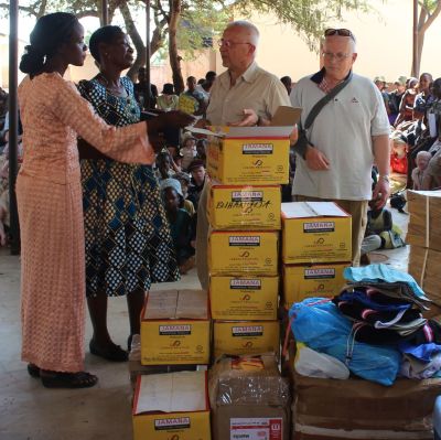 Två (vita) män står vid en stor hög med lådor och andra förpackningar tillsammans med några afrikanska kvinnor. I bakgrunden en massa barn som sitter och följer med. Platsen är i en skola i Tanzania i Afrika.