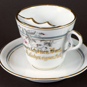 Skäggmugg (kring 1890) Barttasse mit der Aufschrift „Zuschonen Deinen schönen Bart / Nimm dieses Tässchen eigner Art.“. KPM Berlin, um 1890. Porzellan, bemalt.