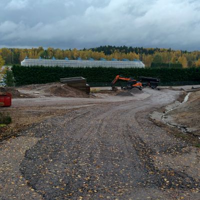 Rinteeseen maansiirtotöinä rakennettua kivistä latupohjaa. Kaivinkone. Keskellä kuvaa rakenteilla olevan kävelysillan betonista valetut päät. Rinteen alla silla takana näkyy stadionin kuusiaita, jonka takaa pilkottaa remontissa oleva huputettu urheilukentän katsomo.