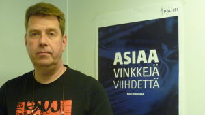 Thomas Ljunggren jobbar vid Raseborgs polisstation. Med tanke på polisens resurser vore det bra om hundar hölls kopplade. Polisen fångar hellre riktiga skurkar än hundar. en man, civilklädd polis, som står invid en polisaffisch. På affischen står Asiaa, vinkkejä, viihdettä.