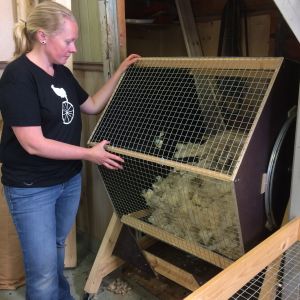 Alicia använder tumlaren som hennes man byggt för att bli av med skräp ur ullen. Kvinna står vid hemmabyggt hjul som innehåller fårull.